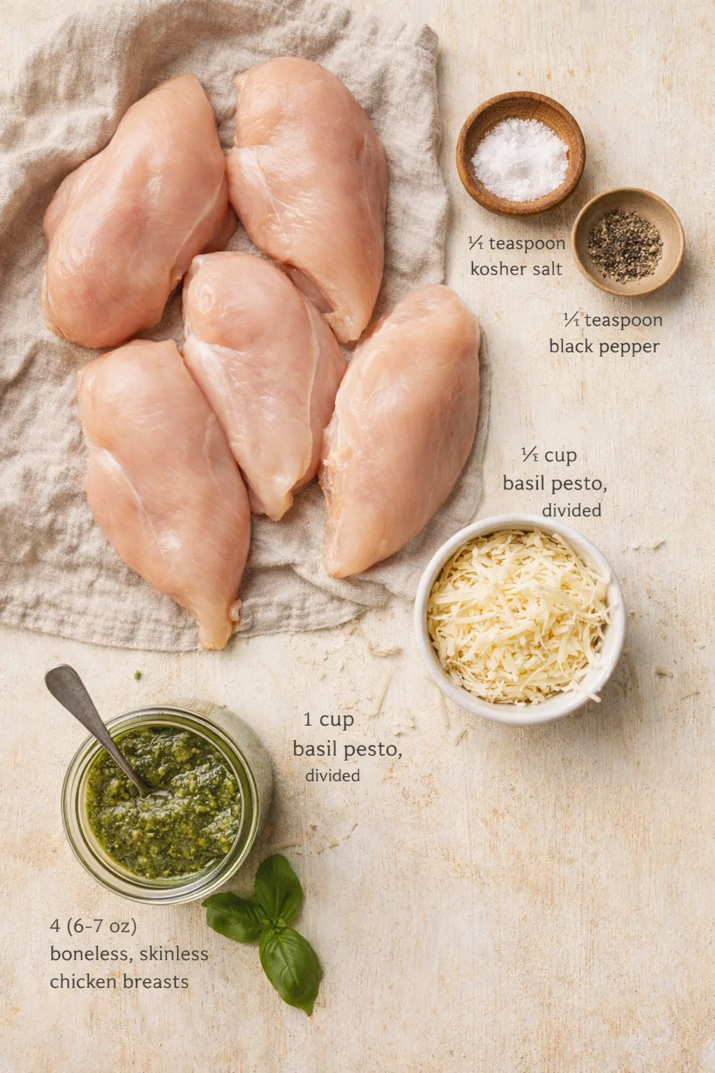 Raw chicken breasts with salt, pepper, pesto, and grated cheese on a light wooden surface.