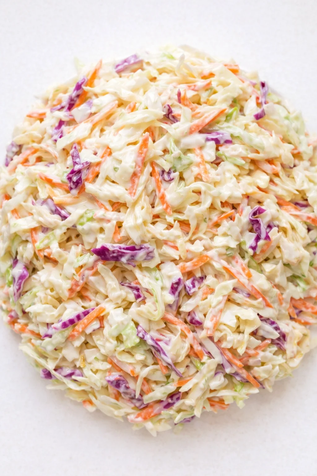 Pile of creamy coleslaw with shredded green and purple cabbage and carrots on a light background.
