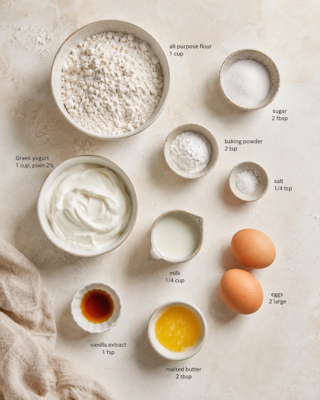 top-down view of baking ingredients arranged on a beige countertop: flour, yogurt, eggs, milk, melted butter, and seasonings.