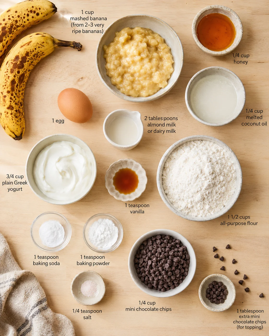 Overhead view of banana-bread ingredients laid out on a wooden surface with labeled bowls.