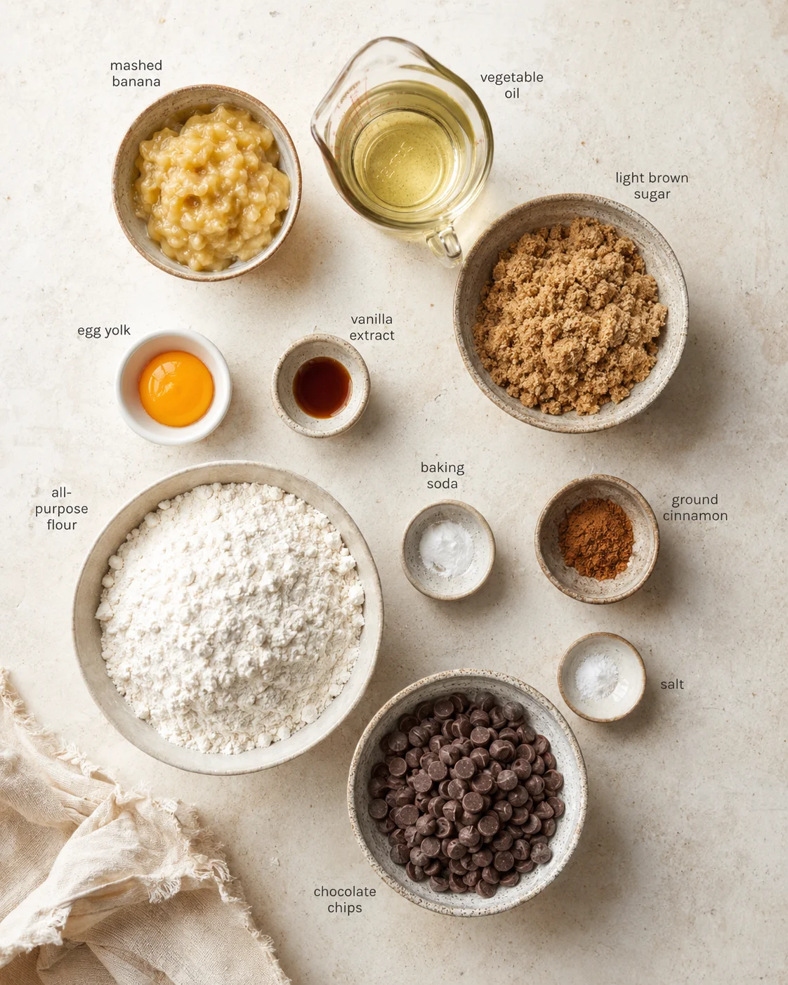 Overhead view of baking ingredients arranged on a light, textured surface with labeled bowls for banana, flour, and chocolate chips.