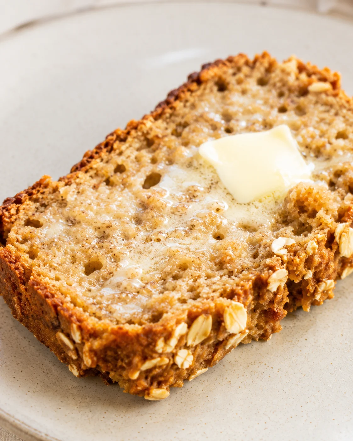 slice of banana-oat bread with a pat of butter on a beige plate