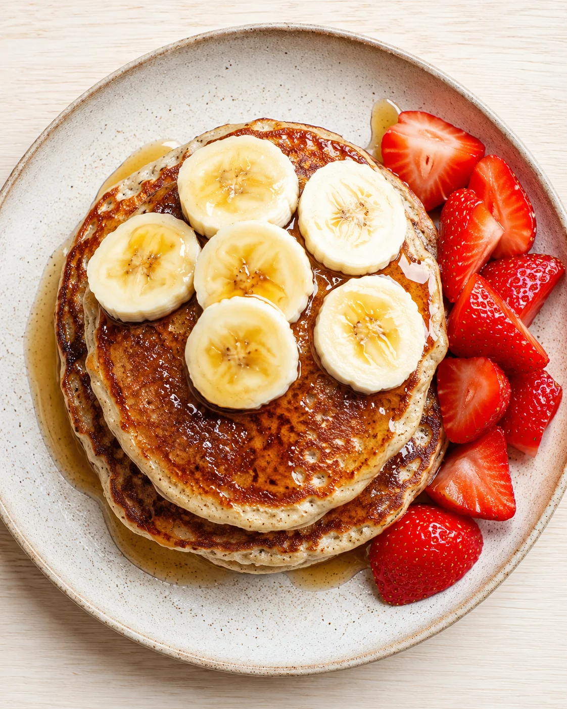 Stack of golden pancakes topped with banana slices, with syrup and fresh strawberries.