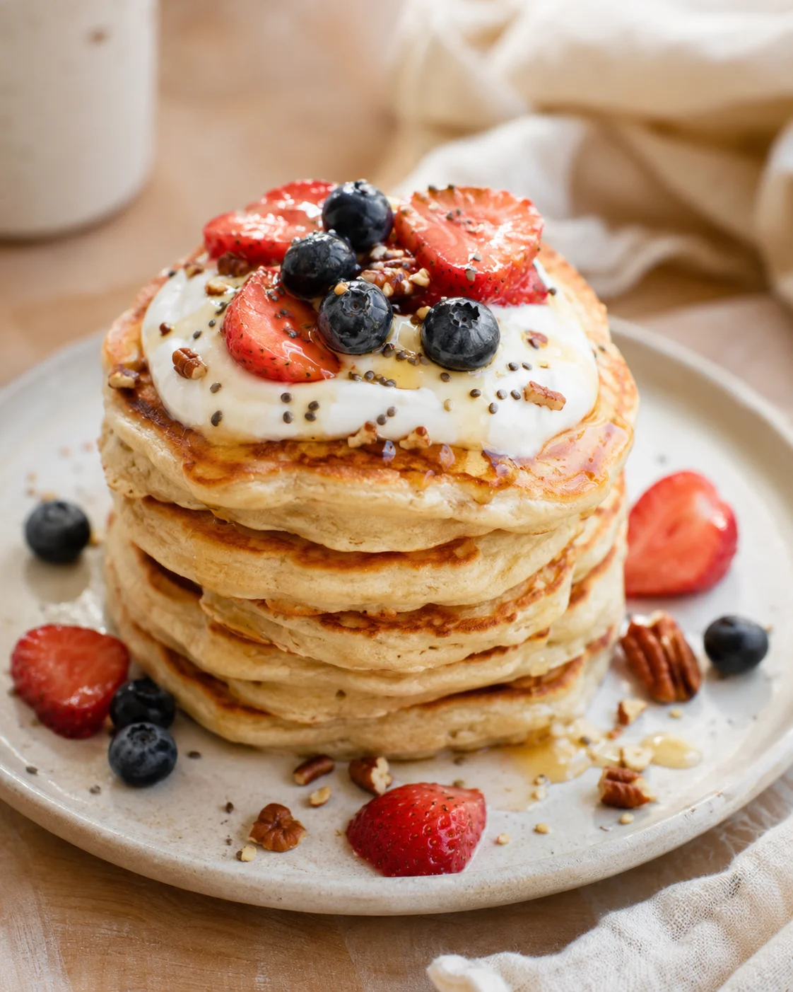 A tall stack of fluffy pancakes topped with whipped cream, strawberries, blueberries, and pecans on a beige plate.