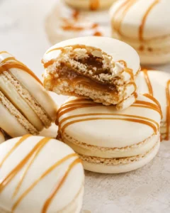 Cream-colored macarons drizzled with caramel, one broken to reveal gooey caramel filling.