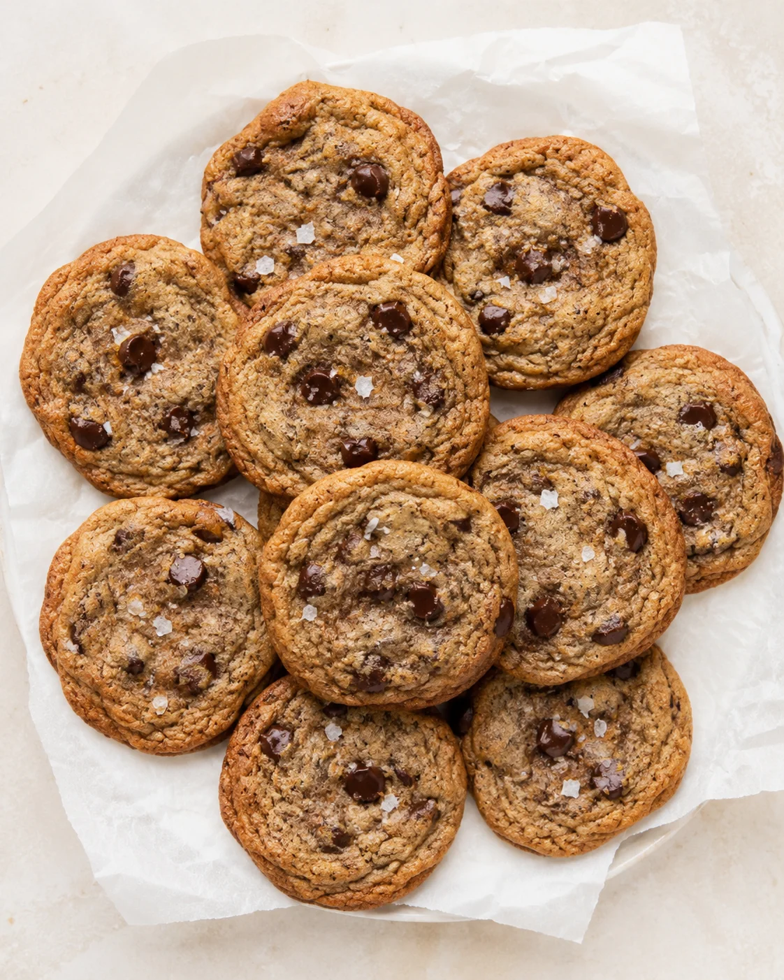 Pile of golden-brown chocolate chip cookies on parchment paper atop a light surface.
