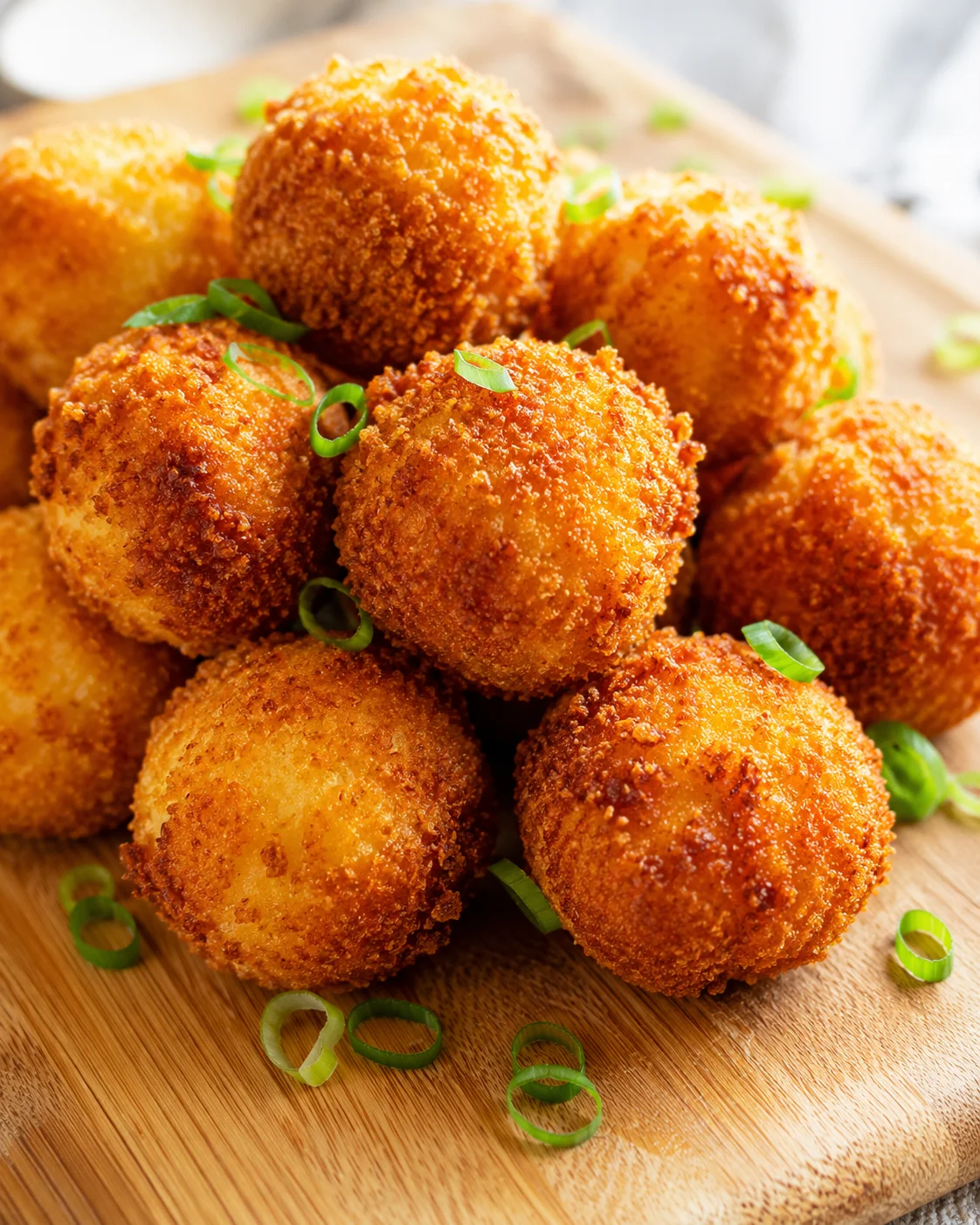 pile of golden-brown crispy croquettes on a wooden board, garnished with green onion rings.