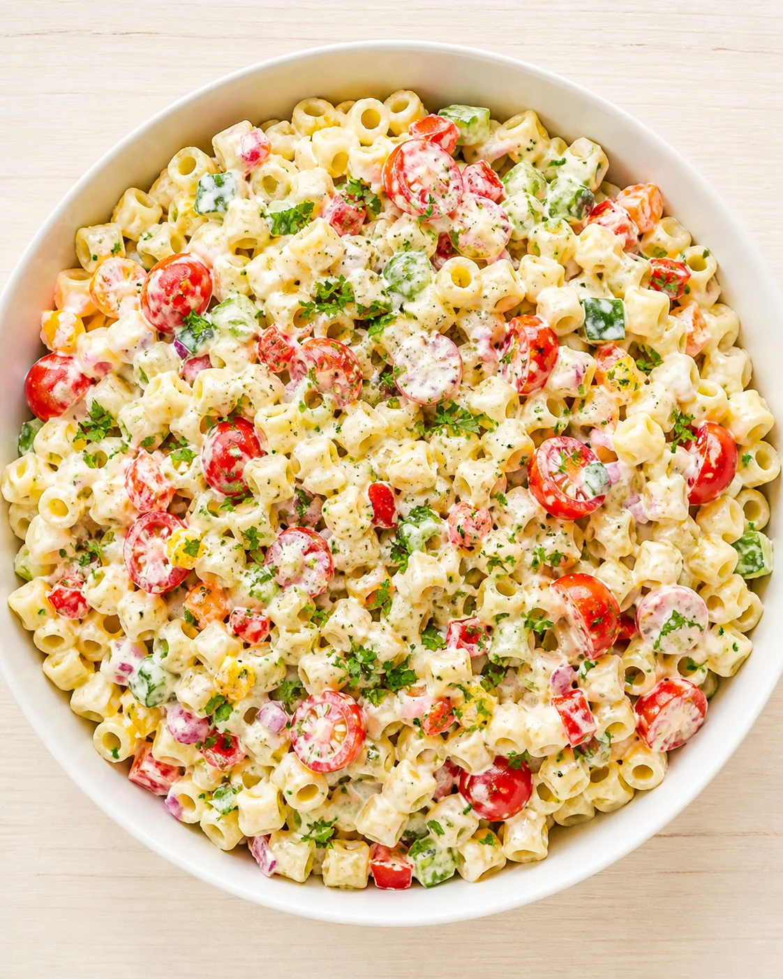 Overhead view of a white bowl filled with creamy pasta salad, cherry tomatoes, cucumber, and parsley.