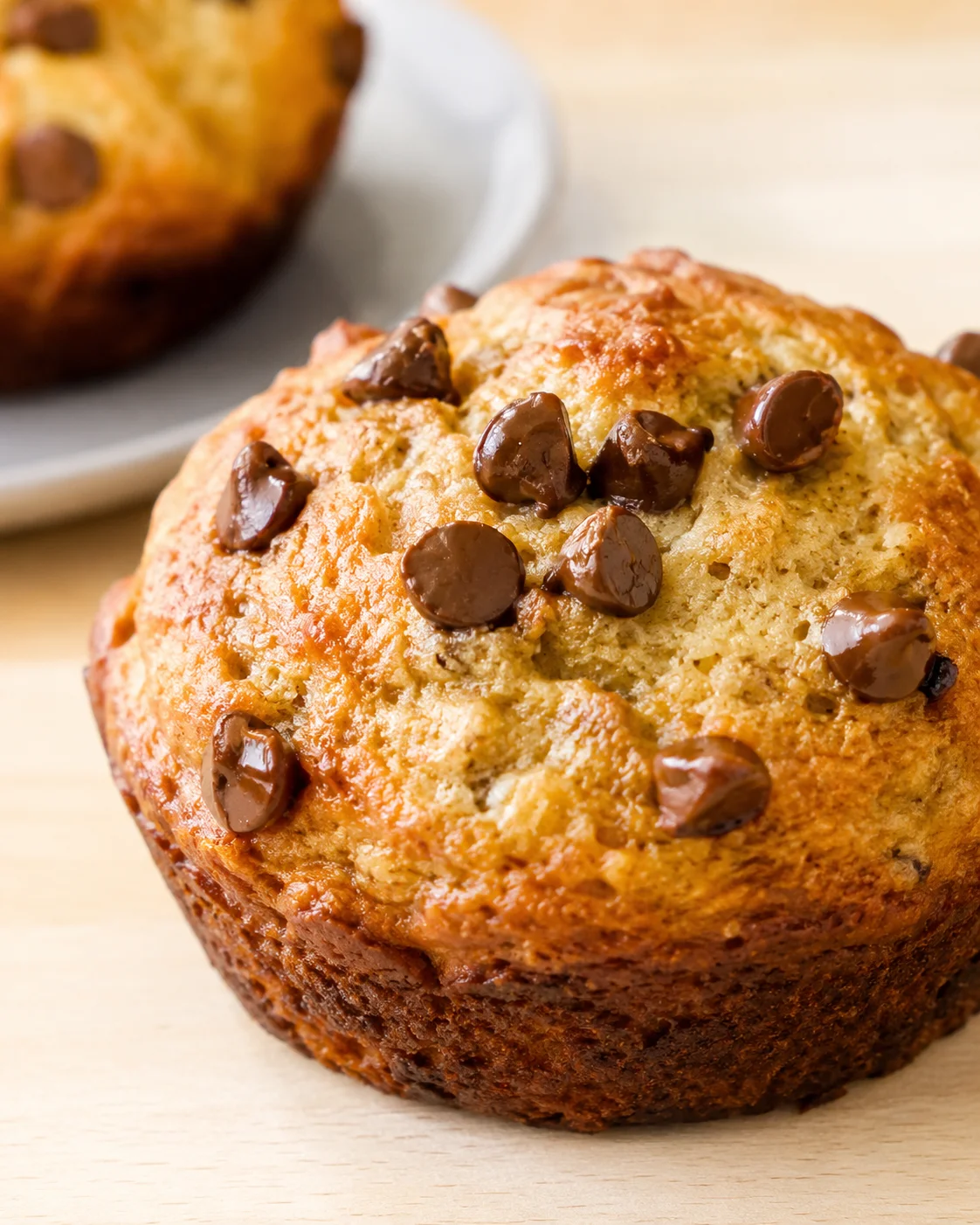 Close-up of a golden-brown chocolate chip muffin on a wooden surface, with another muffin blurred in the background.