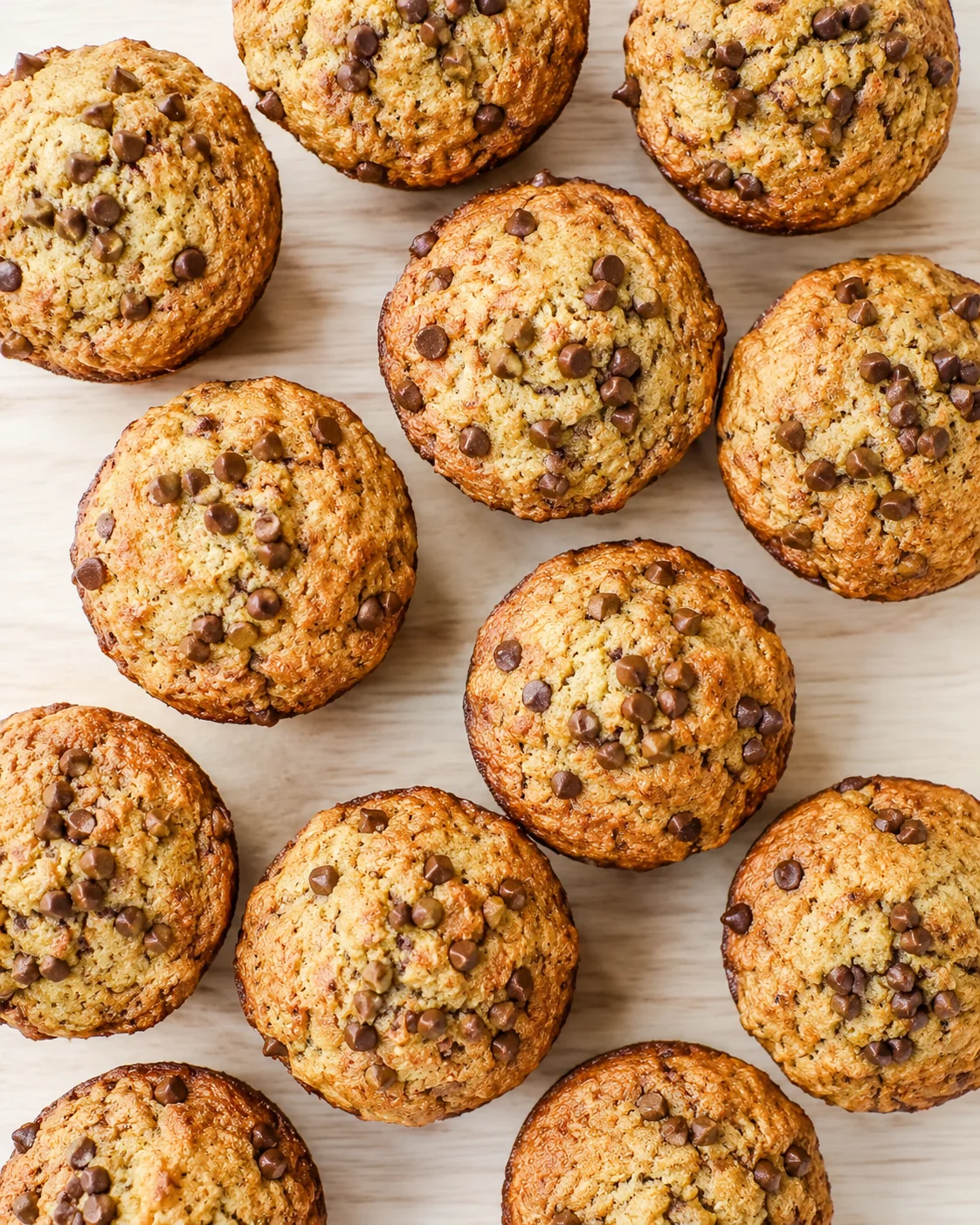 top-down view of golden-brown muffins dotted with chocolate chips on a light wooden surface