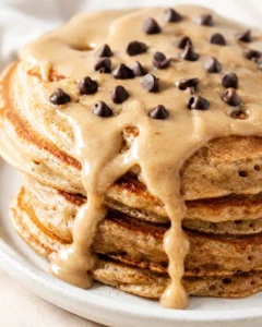 Close-up of a stack of fluffy pancakes drenched in peanut-butter sauce and sprinkled with chocolate chips.