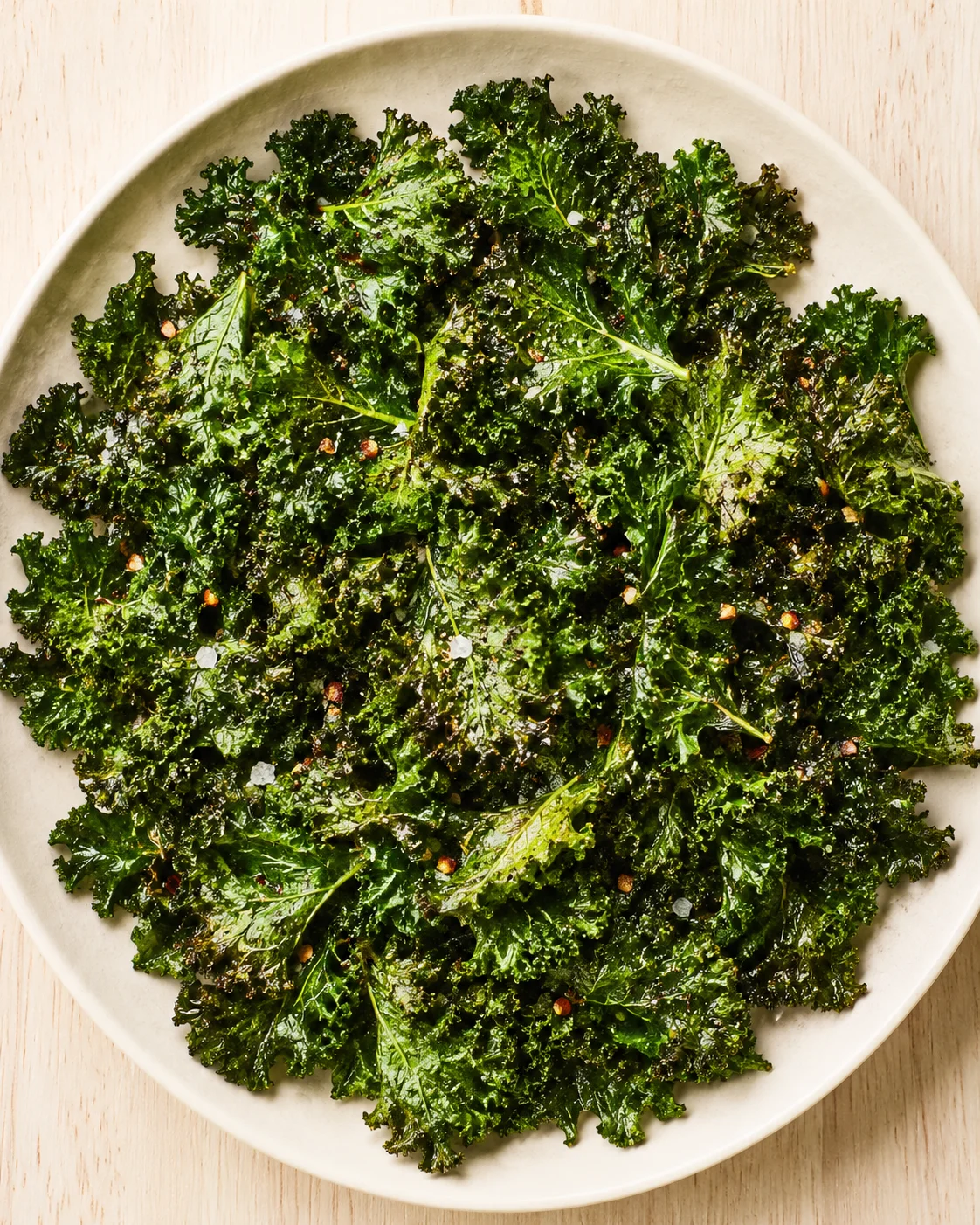 Plate of roasted kale chips with sea salt on a light wooden surface.