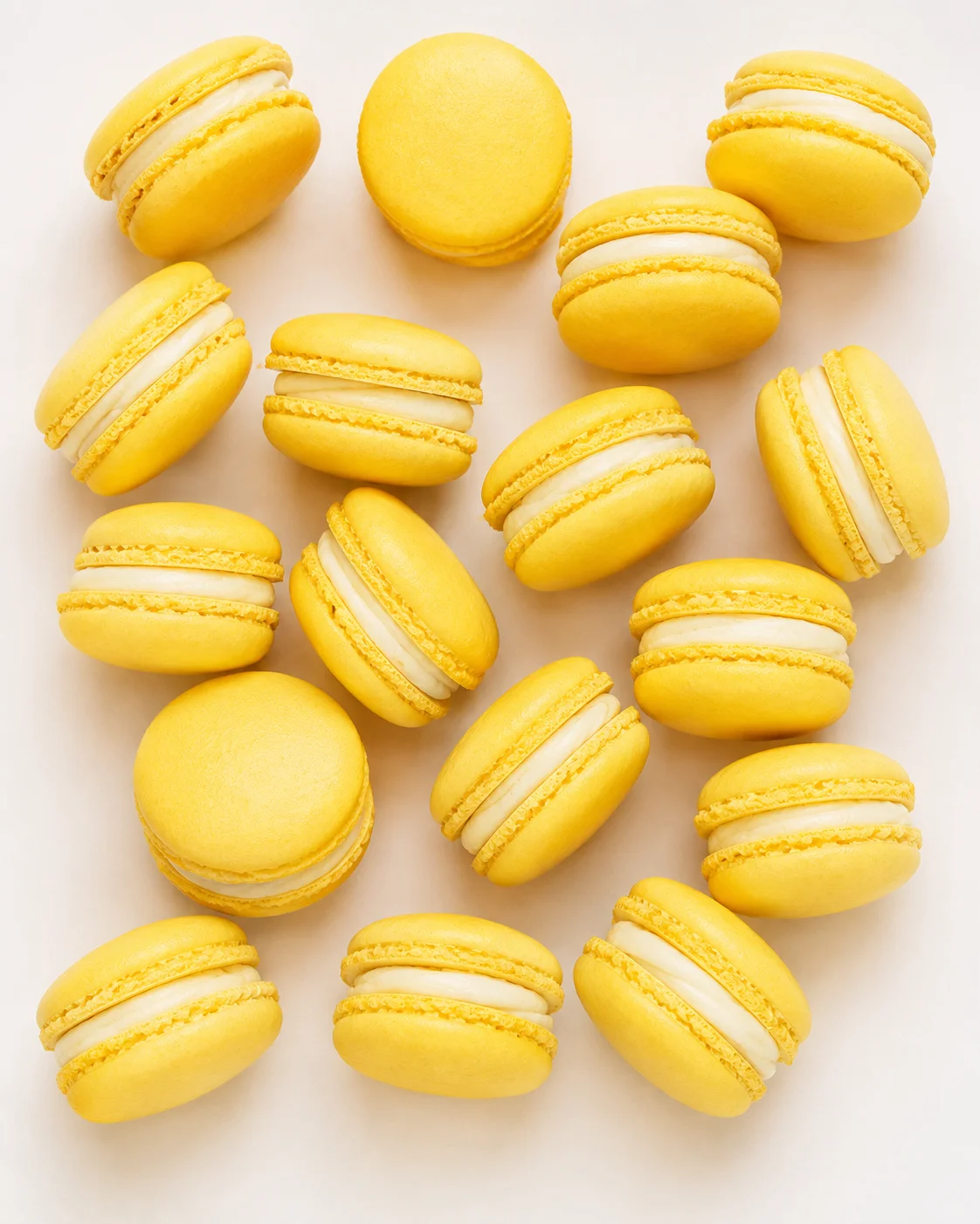 Several yellow macarons with white cream filling scattered on a pale beige background.