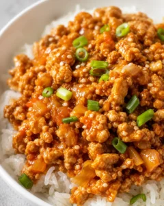 Close-up of vibrant orange ground chicken served over white rice, garnished with green onions.
