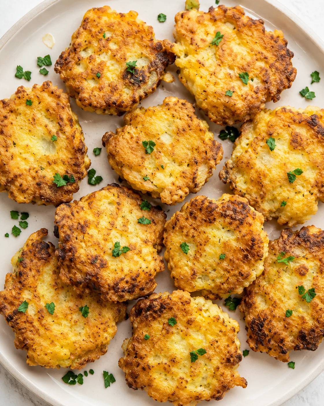 Plate of golden crispy potato fritters sprinkled with chopped parsley on a light ceramic plate.