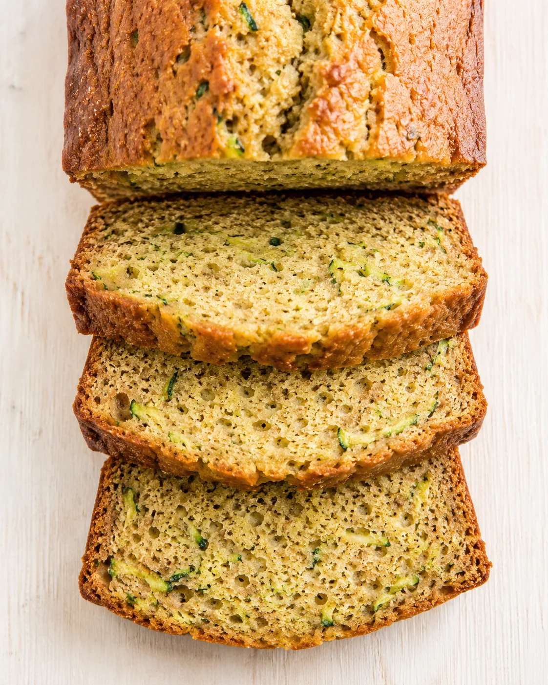 sliced zucchini bread with green specks and a golden crust on a light wooden surface.