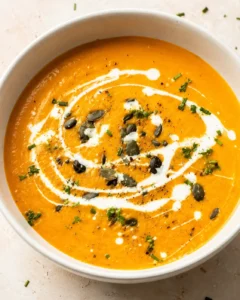 A bowl of creamy roasted carrot and ginger soup garnished with seeds and herbs.