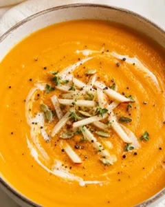 A bowl of vibrant orange carrot and apple soup garnished with thyme, seeds, and cream.