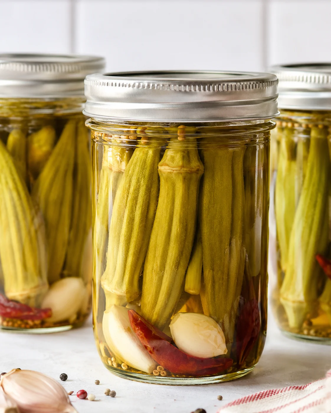 Jars of crunchy pickled okra with garlic and spices on a white surface.