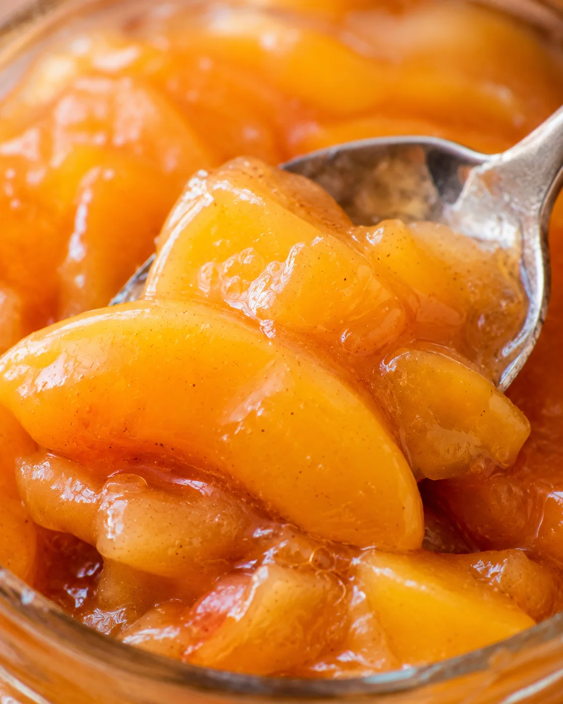 Close-up of homemade peach pie filling in a jar with a spoon, showcasing vibrant peach slices.