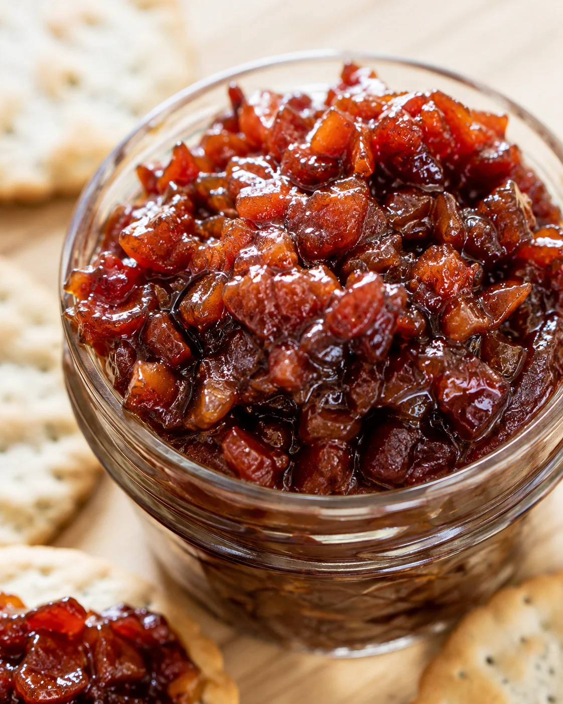 Close-up of flavorful whiskey bacon jam in a glass jar with crackers nearby.