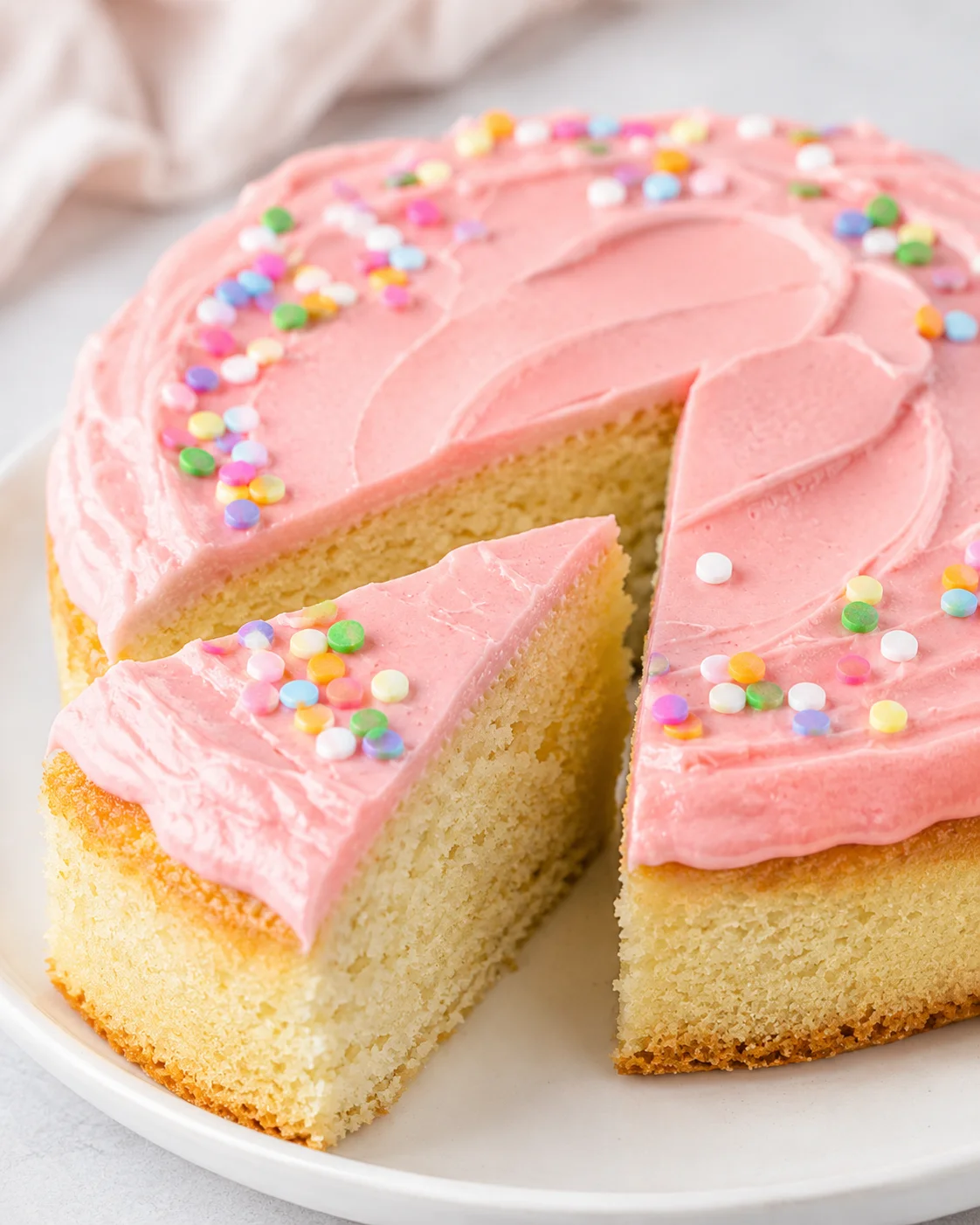 A slice of mini vanilla cake with pink frosting and colorful sprinkles on a white plate