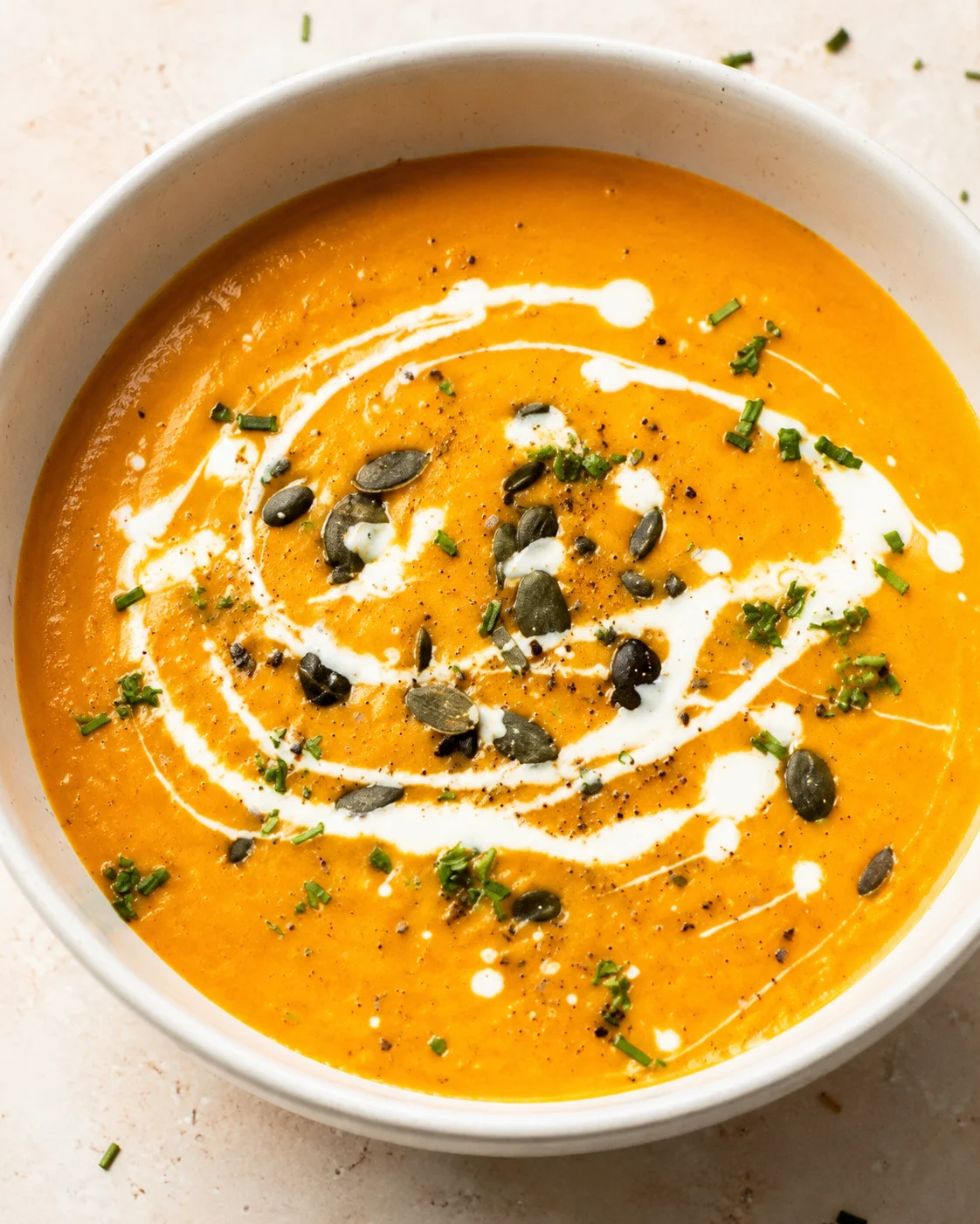 A bowl of creamy roasted carrot and ginger soup garnished with seeds and herbs.