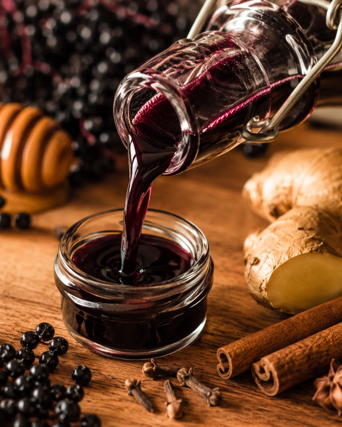 Elderberry syrup being poured from a bottle into a small glass jar with ginger and cinnamon nearby