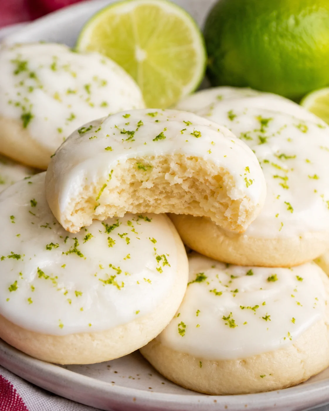 Plate of key lime cookies with one cookie bitten to reveal a soft interior, garnished with lime zest and a halved lime in the background