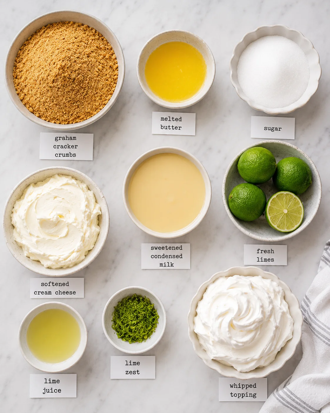 Top-down arrangement of ingredients for no-bake key lime pie with labeled bowls of crumbs, butter, sugar, cream cheese, condensed milk, limes, lime zest and a m