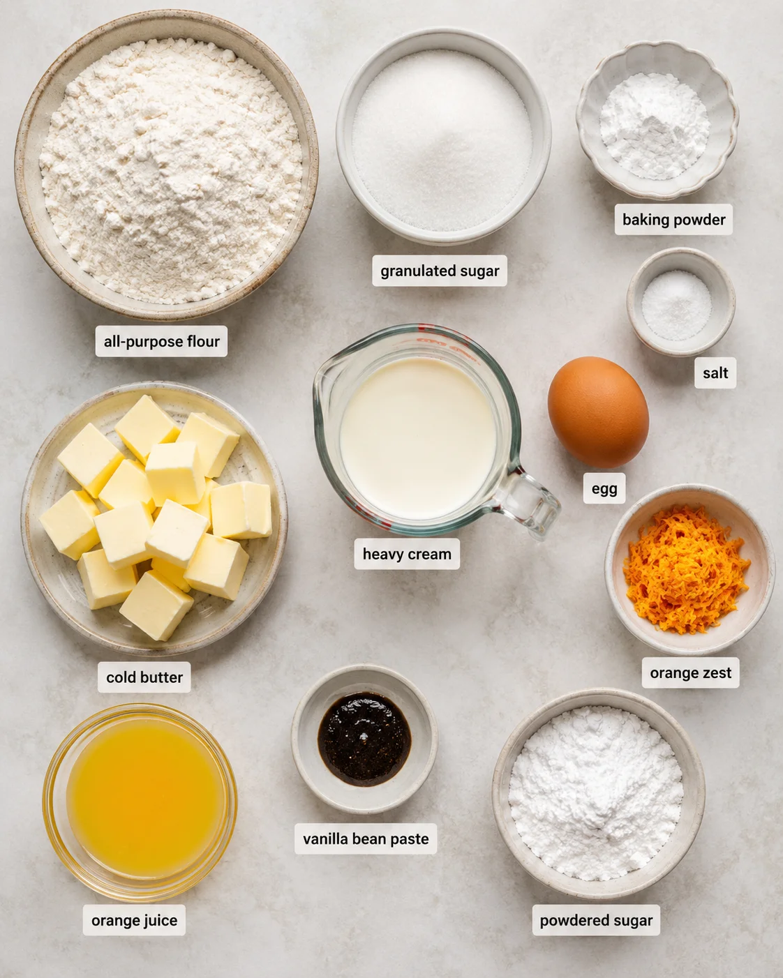 Top-down layout of bowls with flour, sugar, butter, cream, orange zest and juice